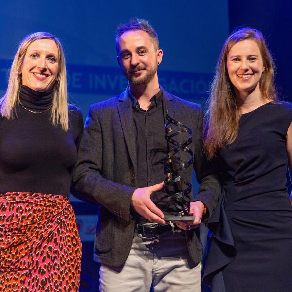 A woman, a man and another woman stand facing the camera, the man in the middle holds a metal award- a sculpture of two film strips in a double helix.